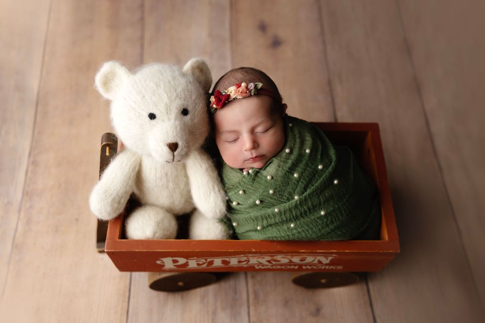 Sleeping baby in green wrap sharing wooden wagon with white teddy bear.
