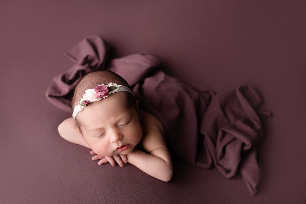 Sleeping newborn with floral headband draped in burgundy fabric against dark background.