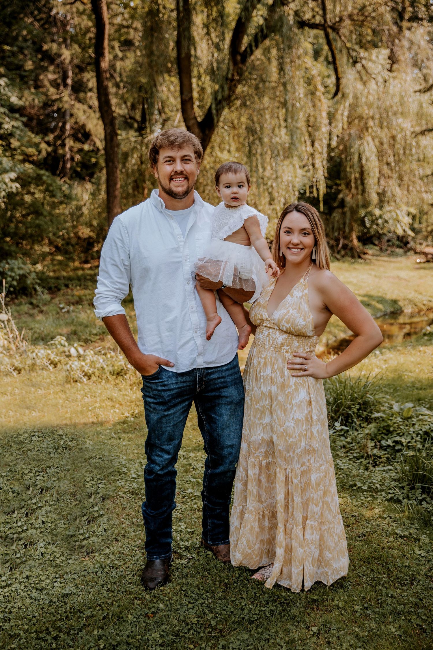 A family poses together in a sunlit forest setting with willow trees in the background.