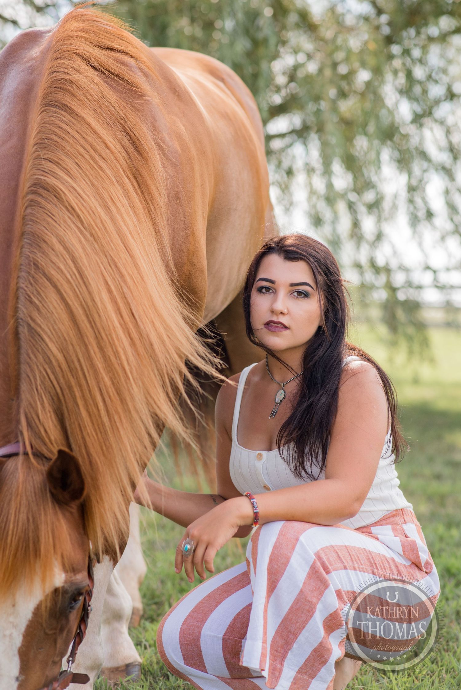 A palomino horse stands next to someone wearing a white top and pink striped pants in a sunny outdoor setting.