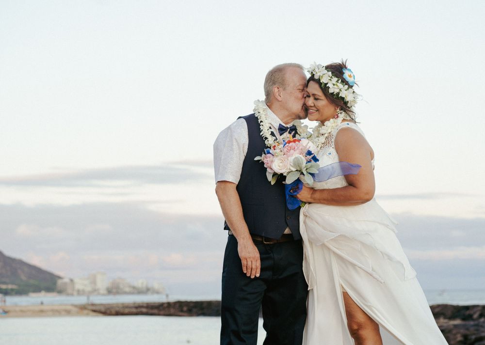 A tender wedding moment captured against coastal backdrop with bride holding floral bouquet.