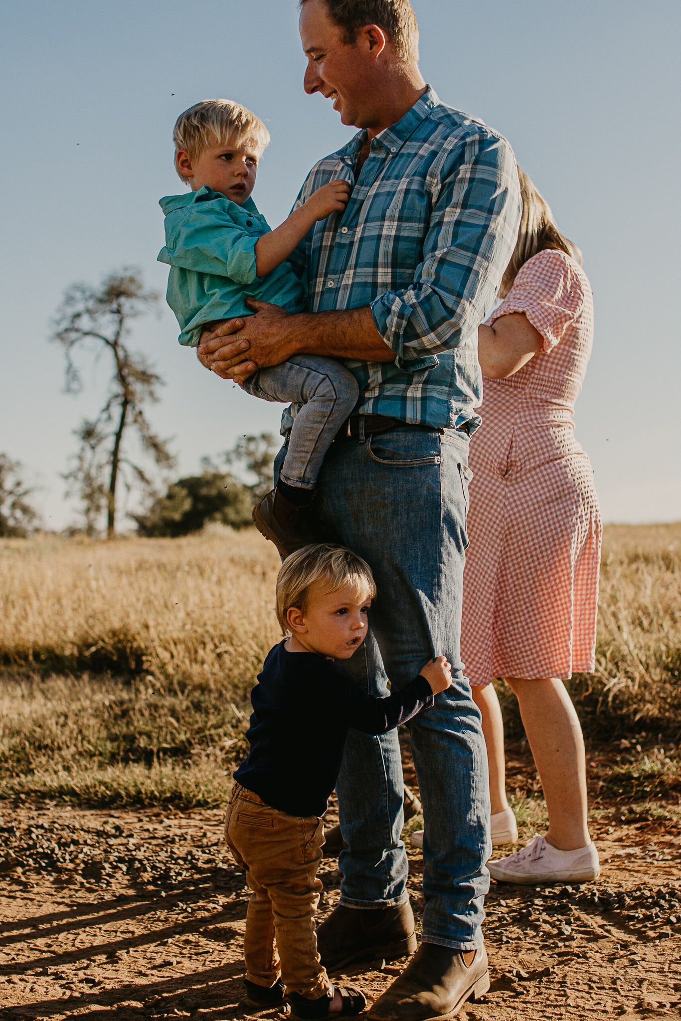 Little boy holding onto Dads legs and Dad holding older boy in a paddock.