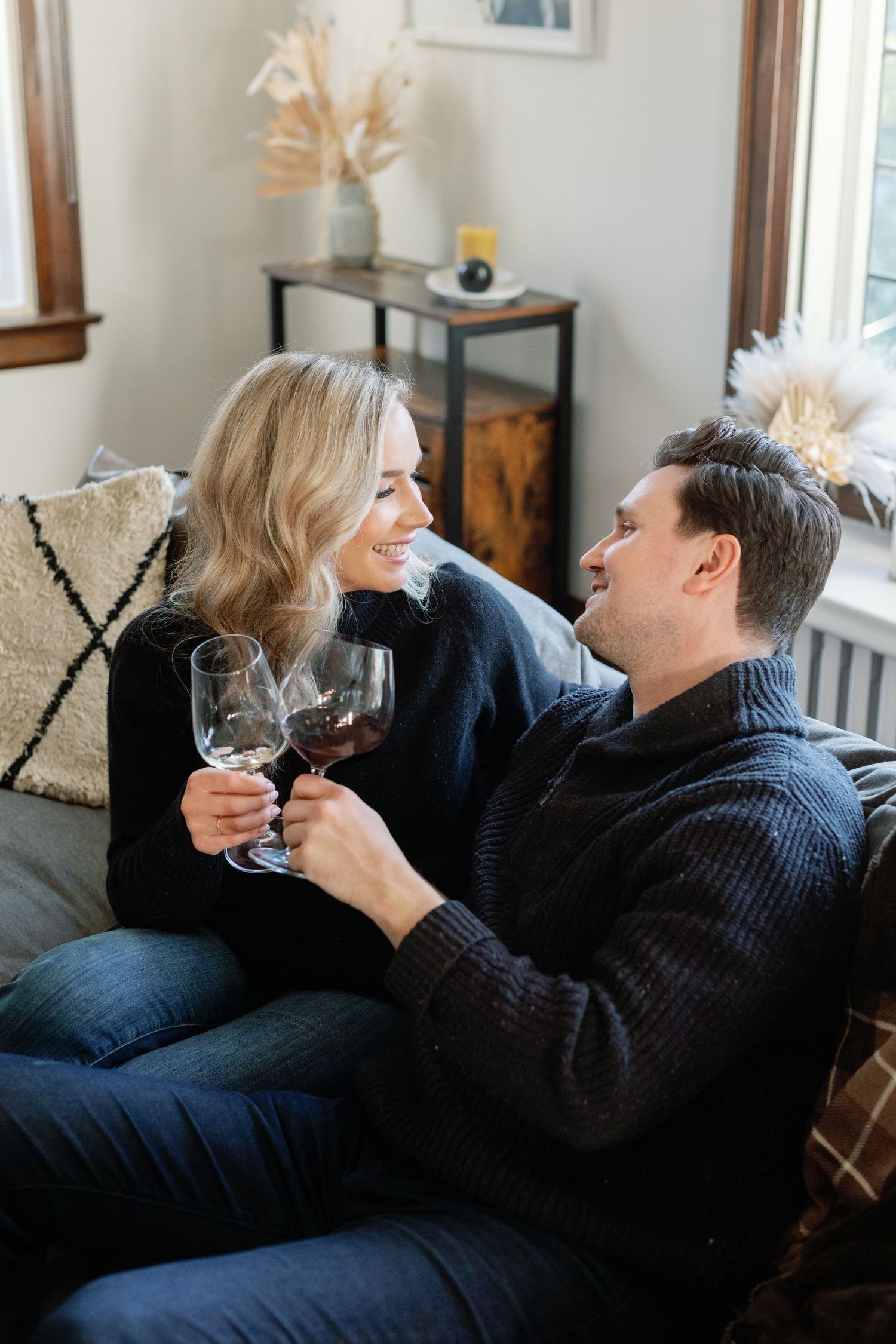 A young couple sit together on a couch smiling and raise wine glasses to each other. Captured by Katie Crane Photography
