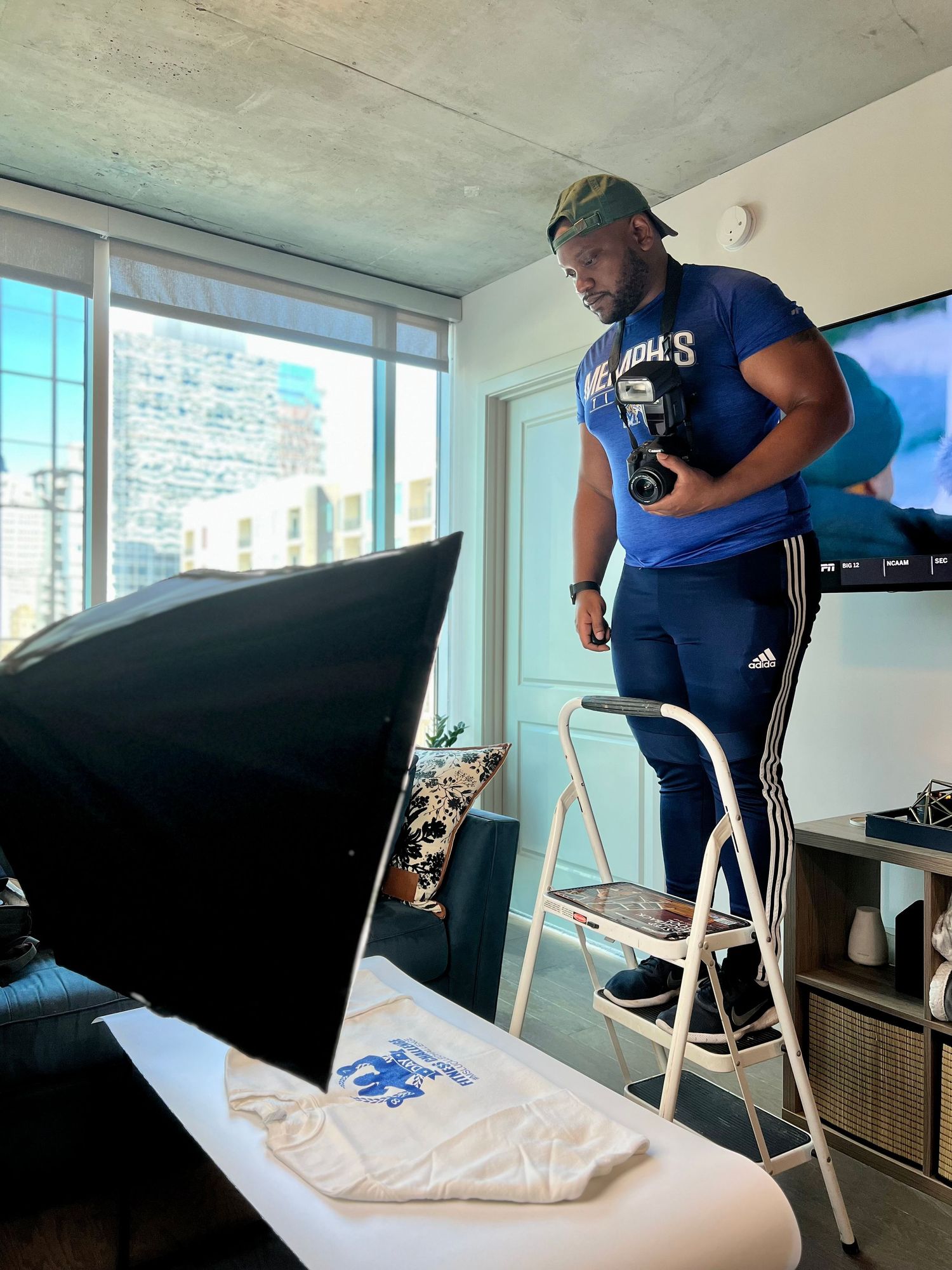 A person in blue athletic wear stands on a ladder during a photoshoot in a modern apartment.