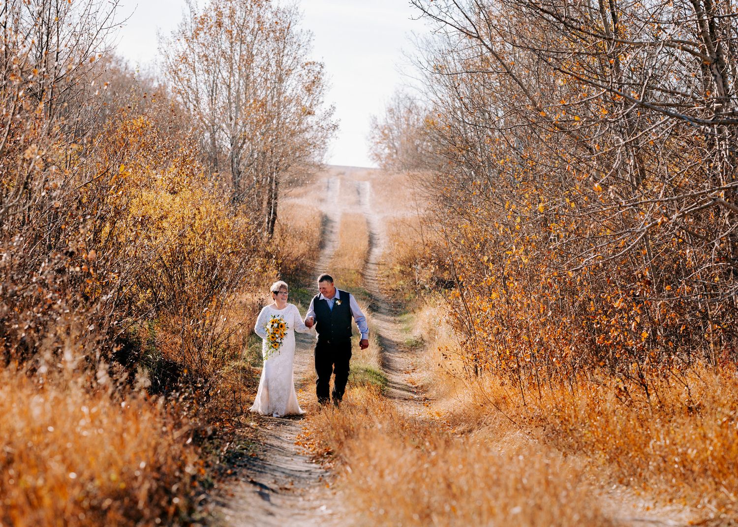 Bride and groom walking hand in hand down a dirt path surrounded by golden autumn trees.