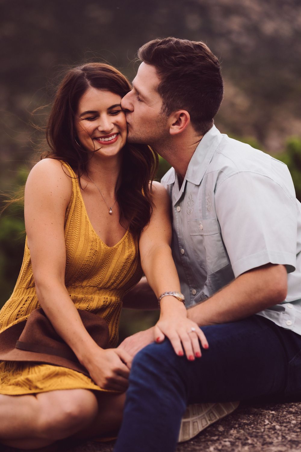 Couple sharing a romantic moment outdoors, woman in yellow dress smiling as man kisses her cheek.