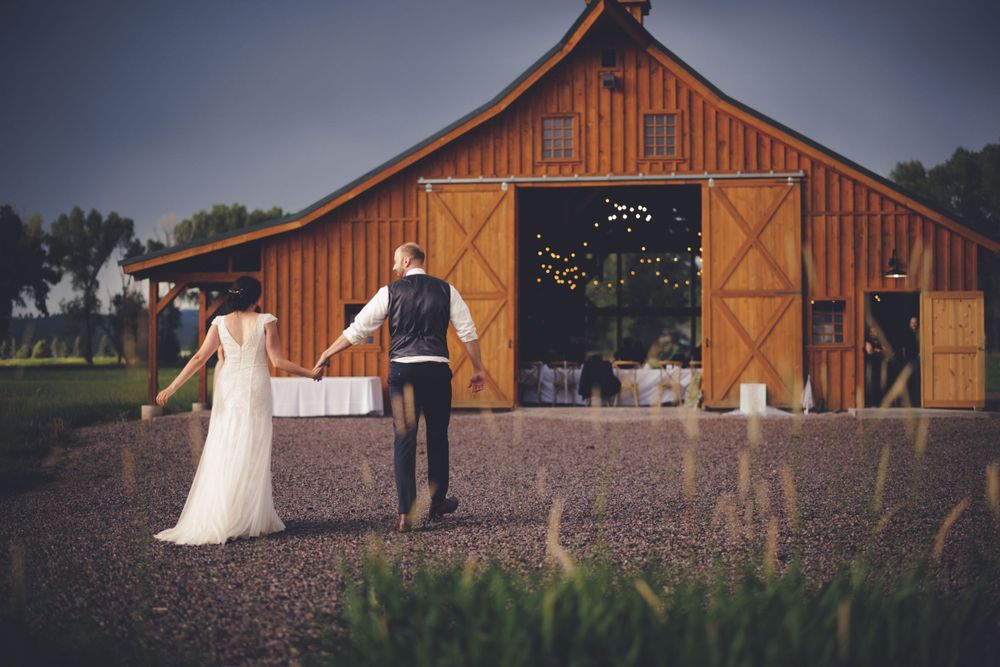 Bride and groom walk hand in hand toward beautifully lit rustic barn wedding venue at dusk.