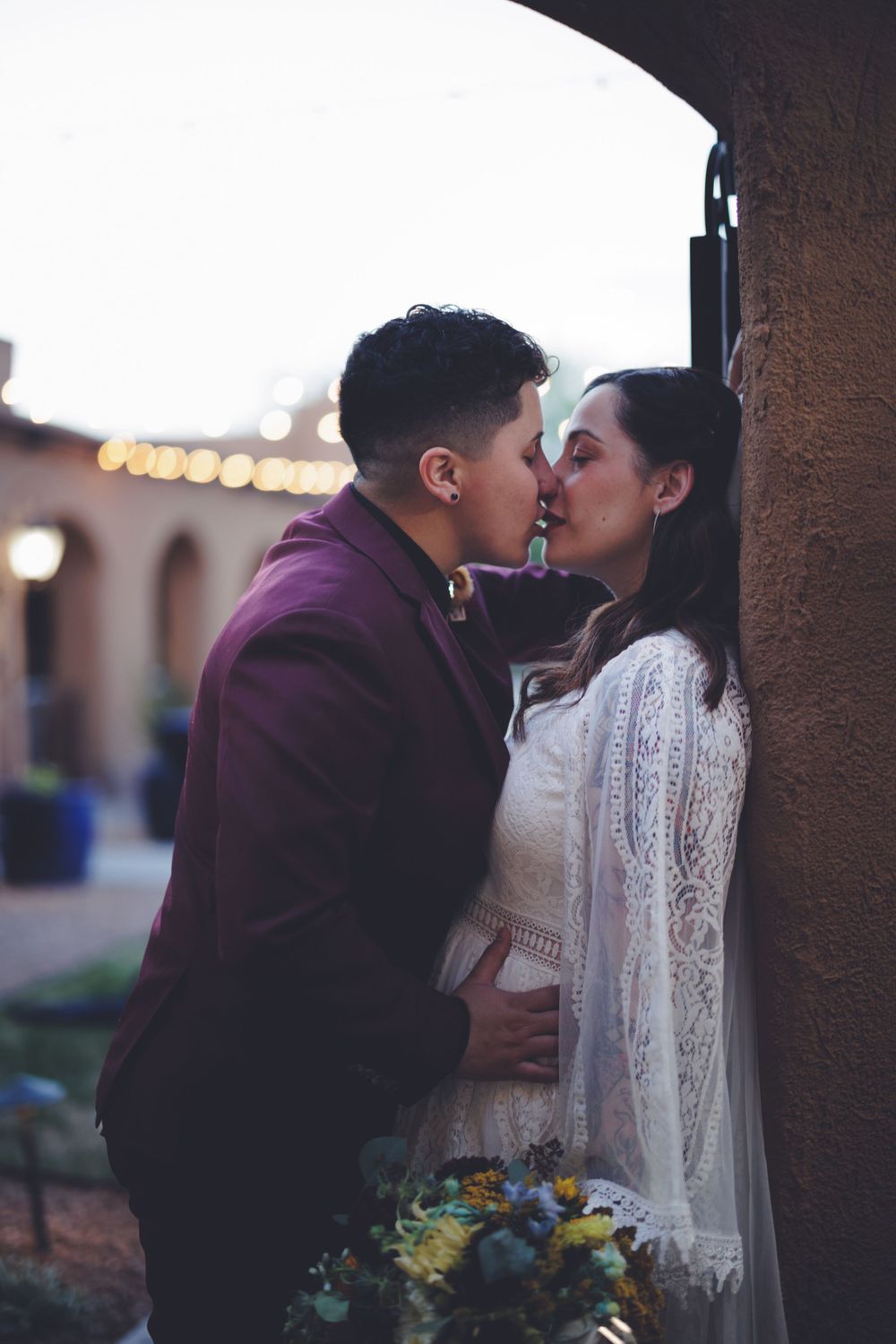 Newlyweds share a romantic kiss beside an archway with bokeh string lights in background.