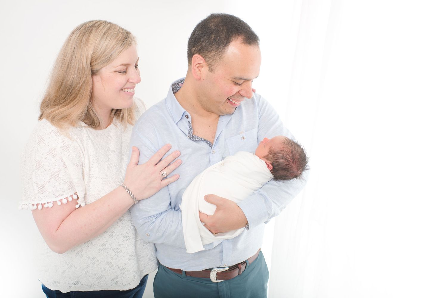 mom wearing a white blouse and dad wearing a blue button down shirt smiling at their newborn baby boy all white studio