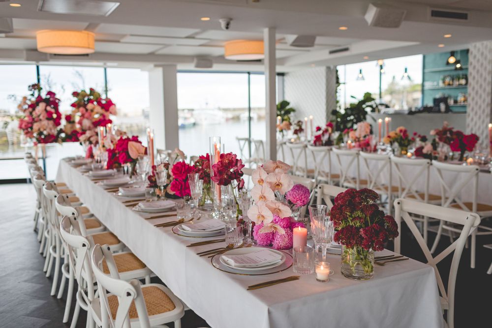 Elegant wedding reception table with pink floral centerpieces and white crossback chairs in a bright venue.