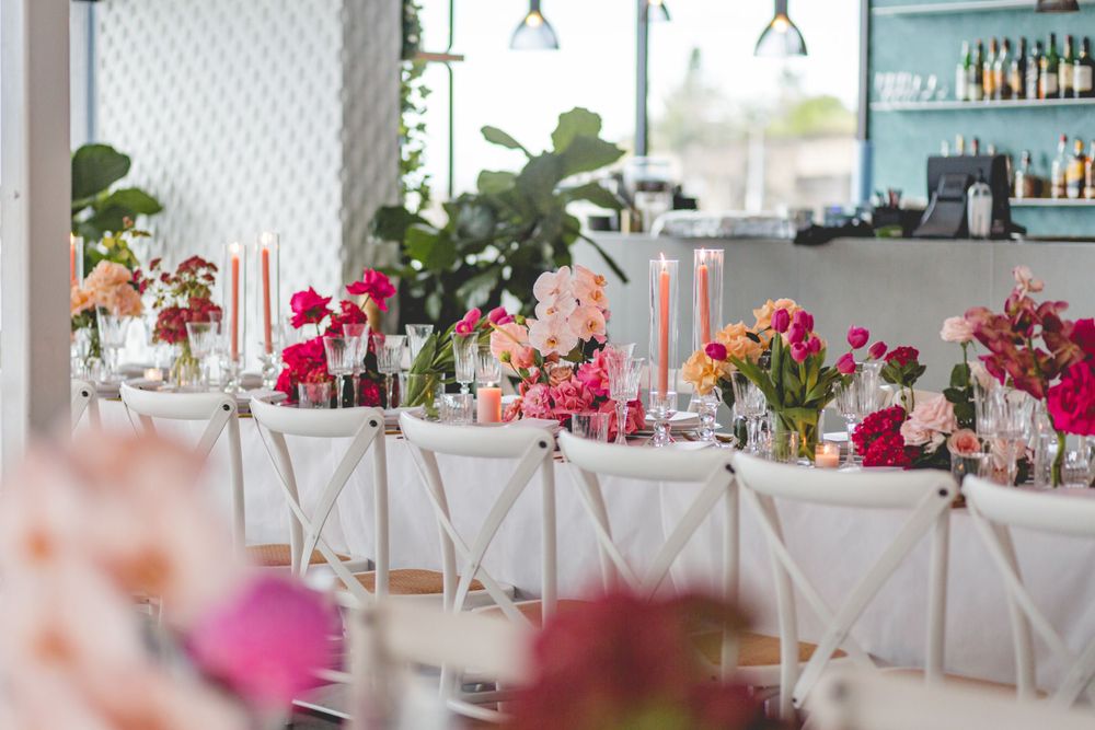Long dining table set with pink floral arrangements and white chairs against a modern bar backdrop.