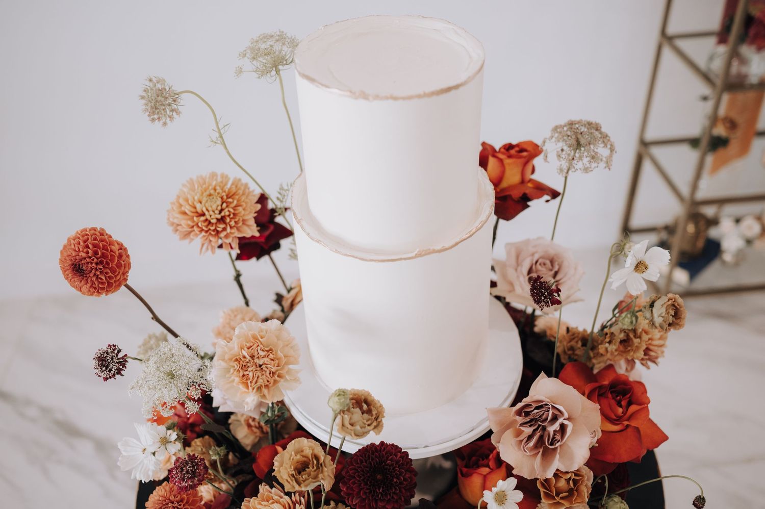 Three-tiered white wedding cake adorned with coral and burgundy flowers against light background.