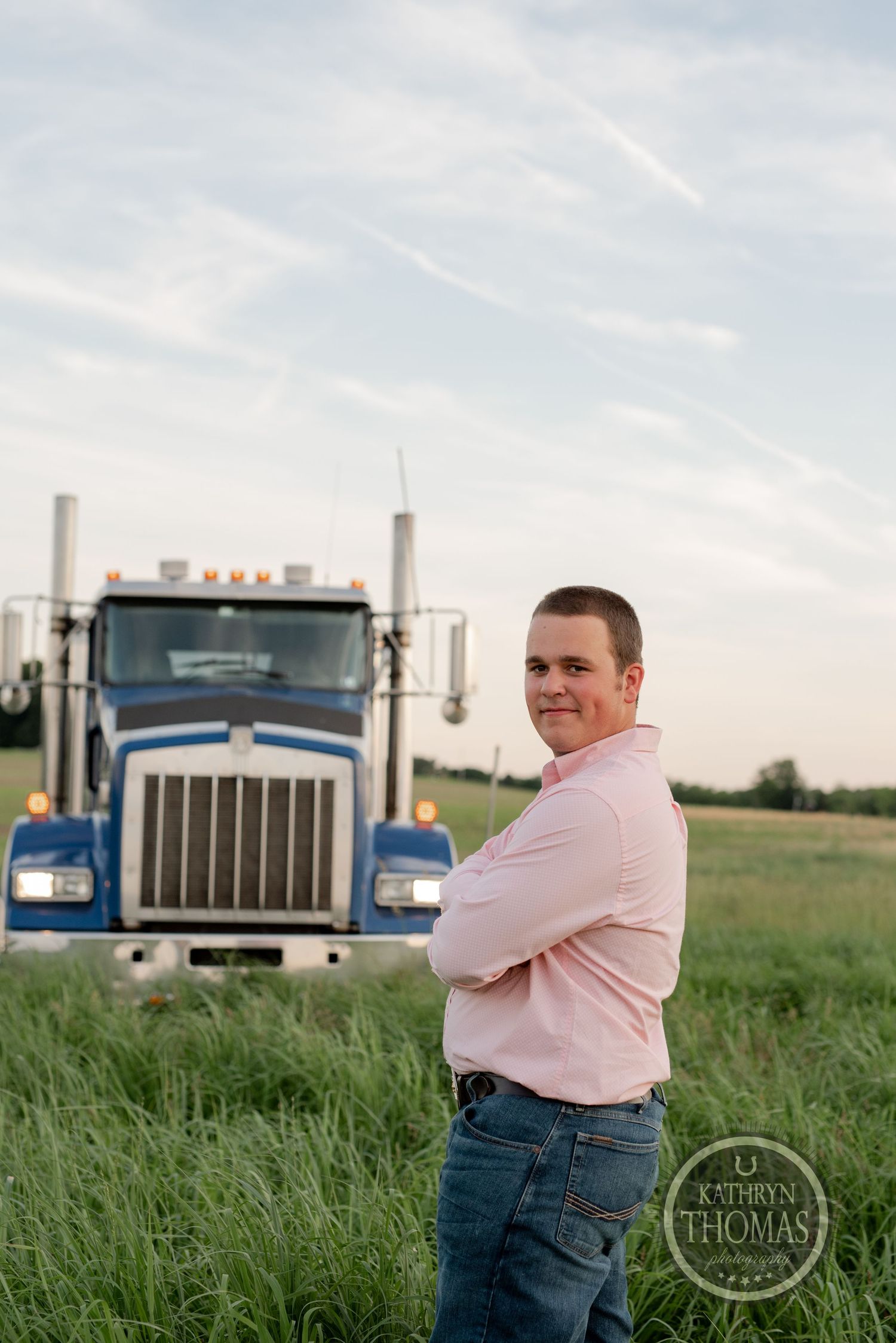Truck driver in pink shirt poses confidently in front of blue Kenworth semi truck at sunset.
