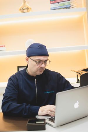 Medical professional wearing scrub cap works on laptop in office setting.