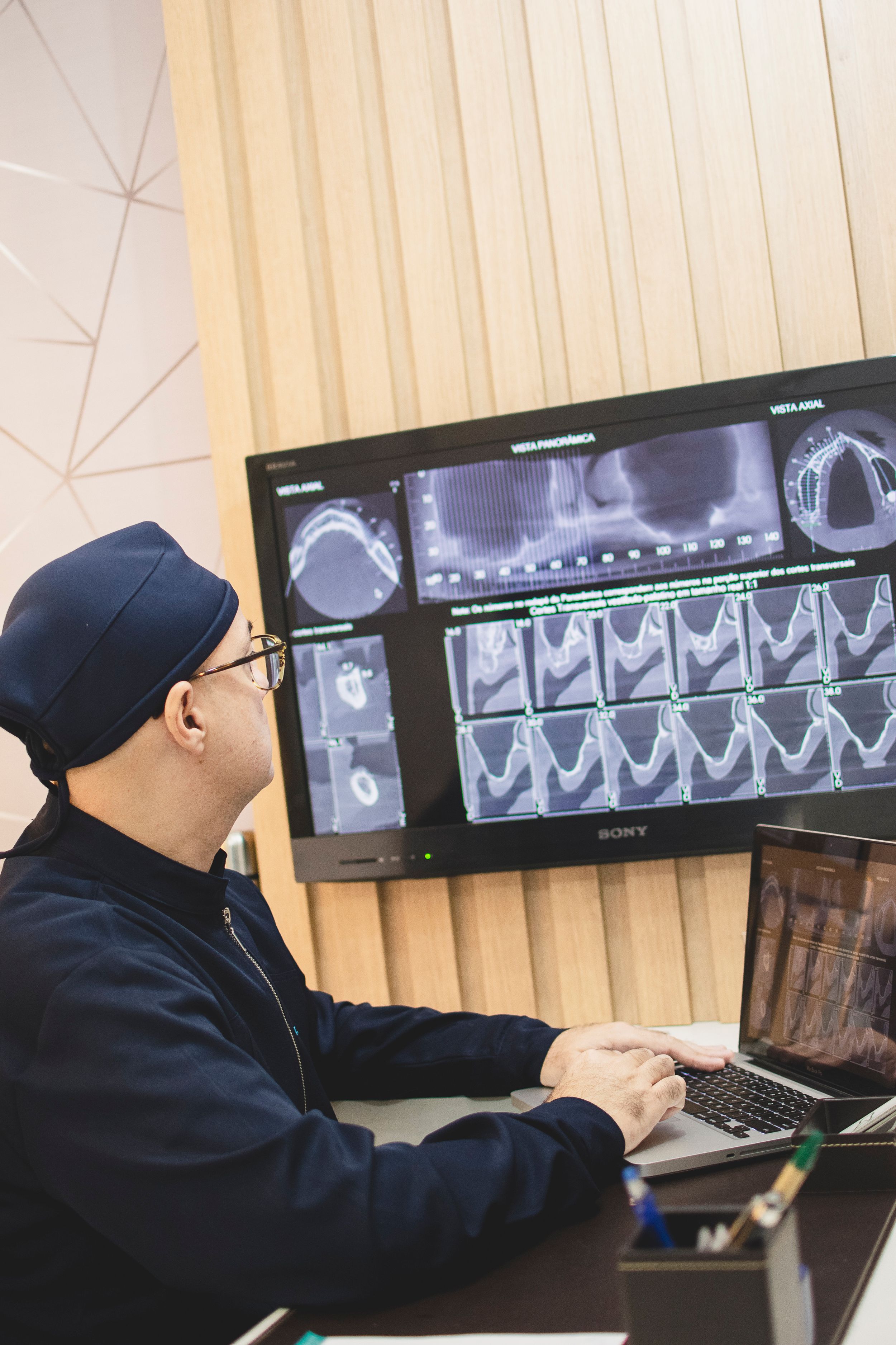 Medical professional in dark clothing examines diagnostic scans on multiple computer monitors.