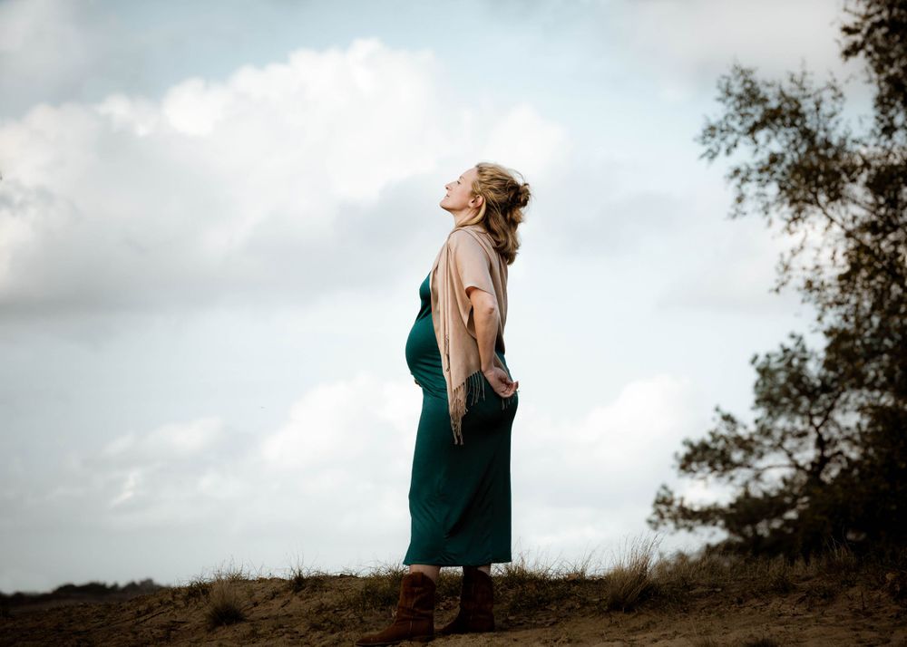 Stralende zwangere vrouw in turquoise jurk kijkt naar de blauwe lucht – zwangerschapsfotoshoot op de Veluwe