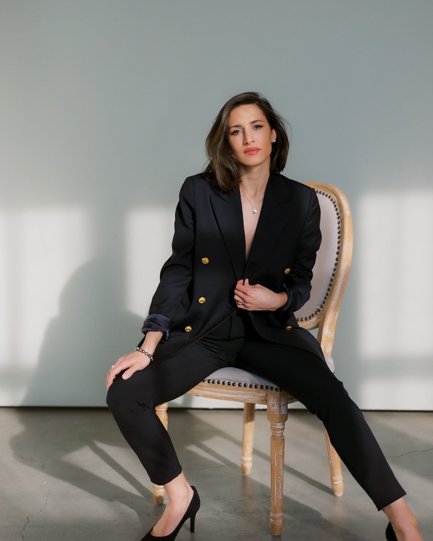 Professional woman in black blazer suit poses on vintage chair in minimalist setting.