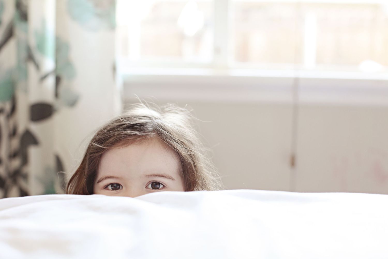 Young child peeking playfully over white bedding in a bright room.