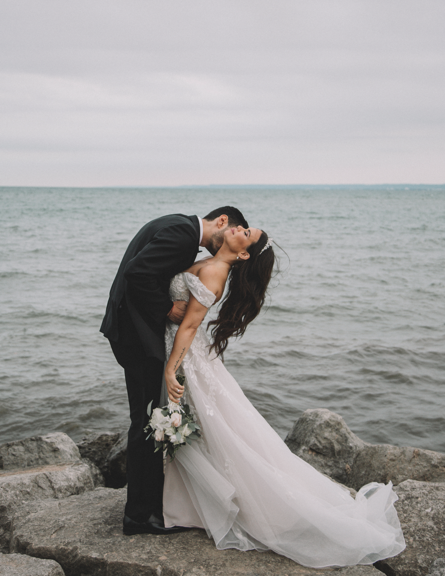 Newlyweds share romantic kiss on rocky shore with ocean waves in background.