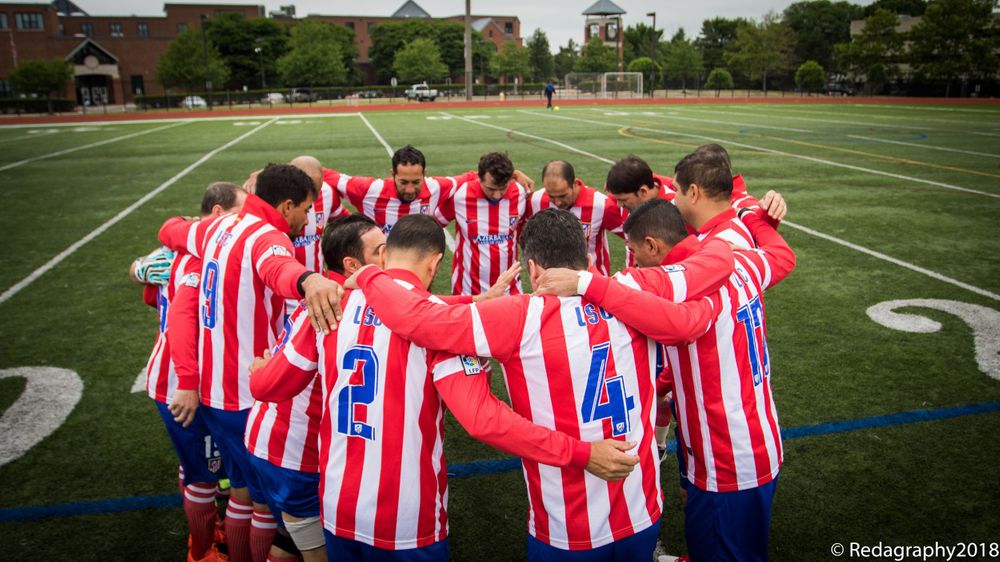 Soccer team in red and white striped uniforms forming a huddle on green turf field.