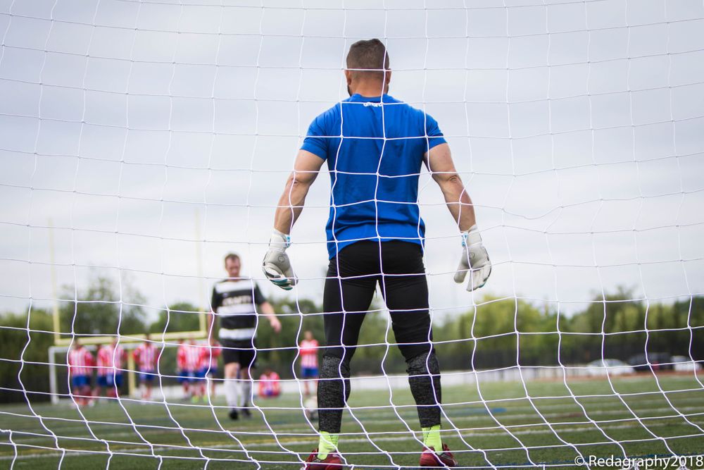 Soccer goalkeeper in blue jersey standing at goal net with gloves ready during outdoor game.