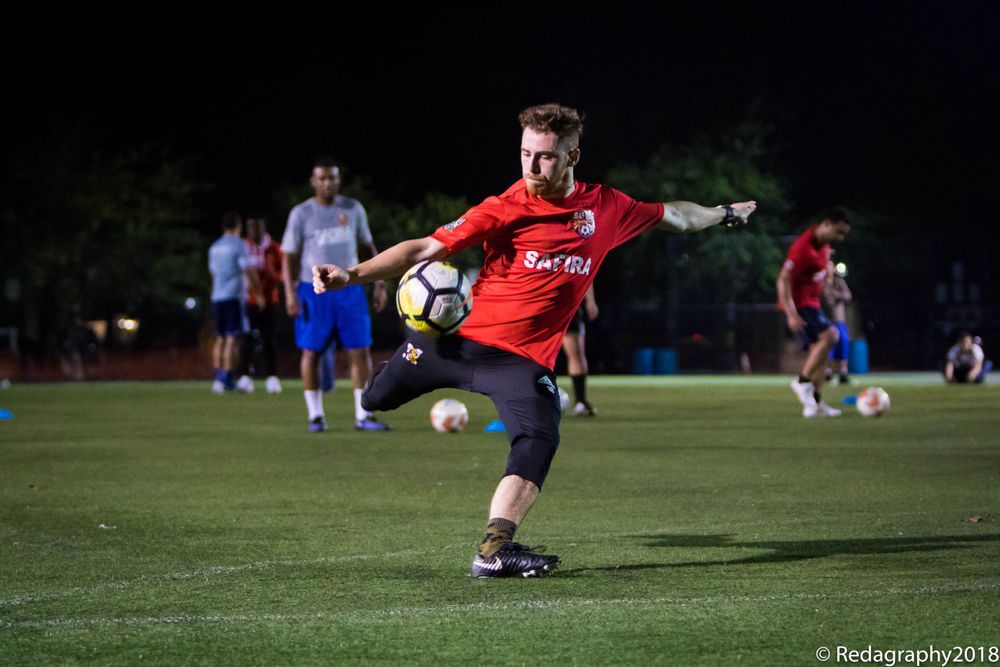 Soccer player in red striped jersey kicking ball during evening practice on illuminated field.