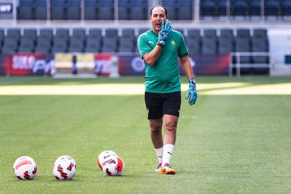 Soccer goalkeeper in green jersey directing players during warm-up with multiple balls on field.