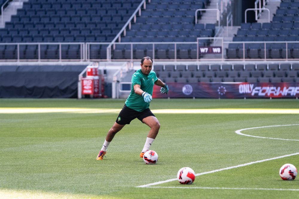 Soccer goalkeeper in green jersey dribbling ball during training session in empty stadium.