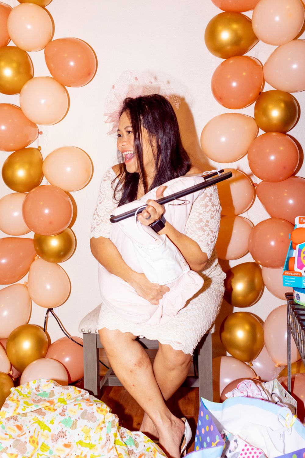 A soon-to-be mother sitting in a chair holding a gift and laughing towards a guest with balloons on both sides of them.