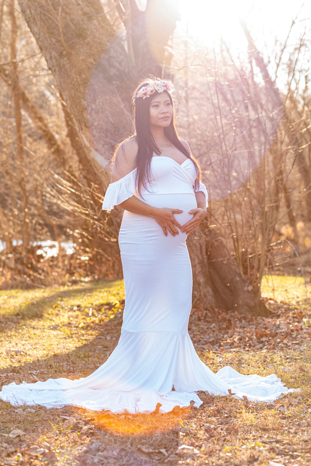 A soon-to-be mother wearing a white dress standing with her hands on her stomach looking out.
