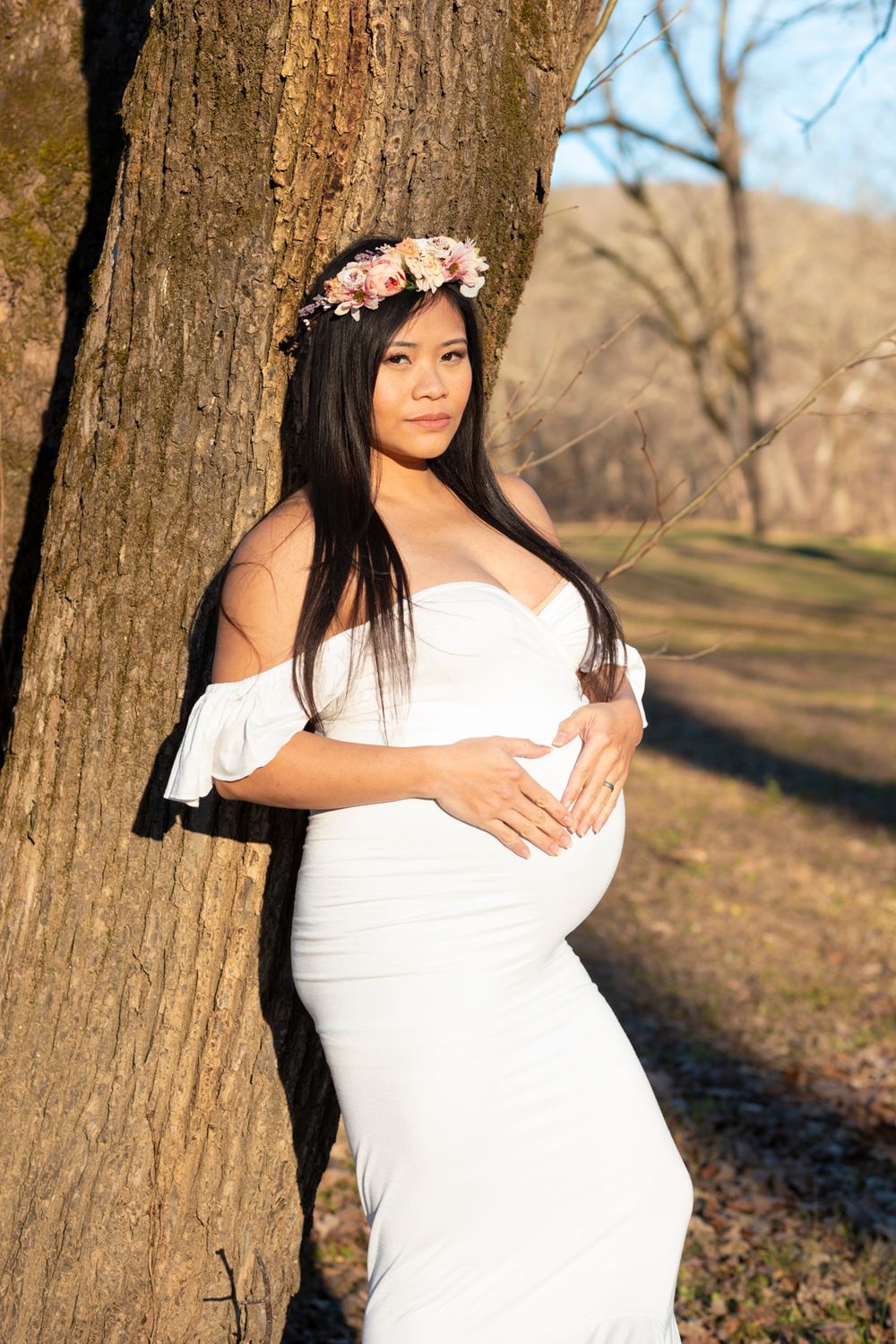 A soon-to-be mother wearing a white dress leaning on a tree with her hands on her stomach in a heart shape.