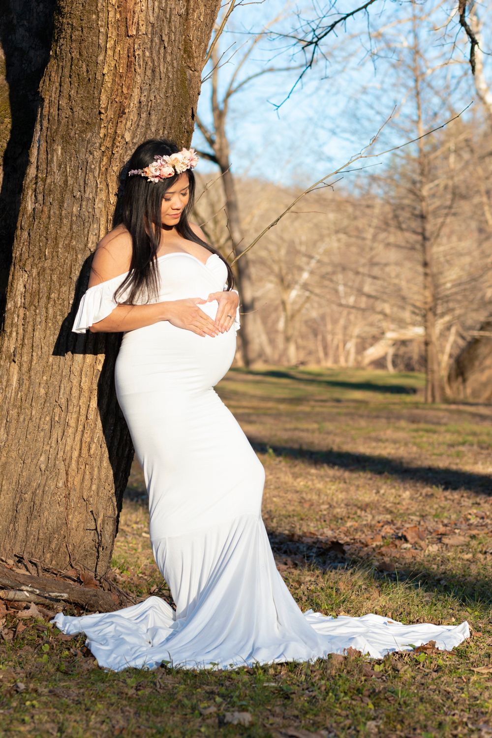 A soon-to-be mother wearing a white dress leaning on a tree looking at her stomach.