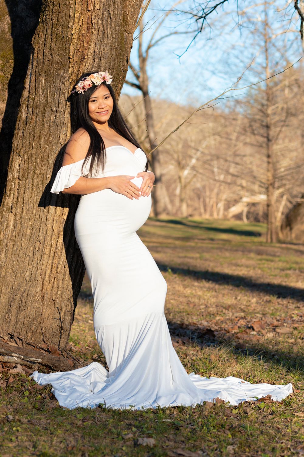 A soon-to-be mother wearing a white dress leaning on a tree with her hands in a heart shape on her stomach.