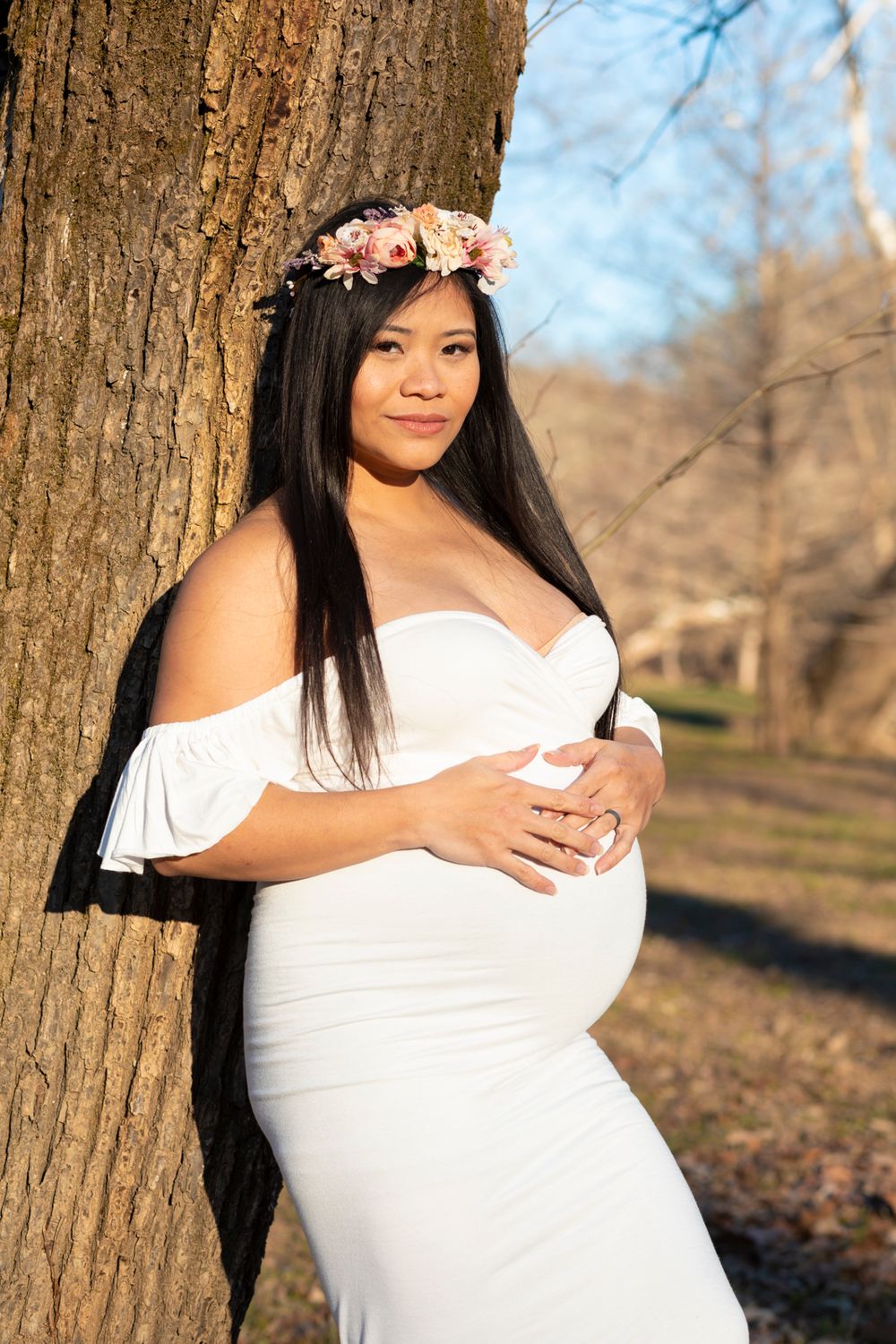 A soon-to-be mother wearing a white dress leaning on a tree with her hands on her stomach looking at the camera.