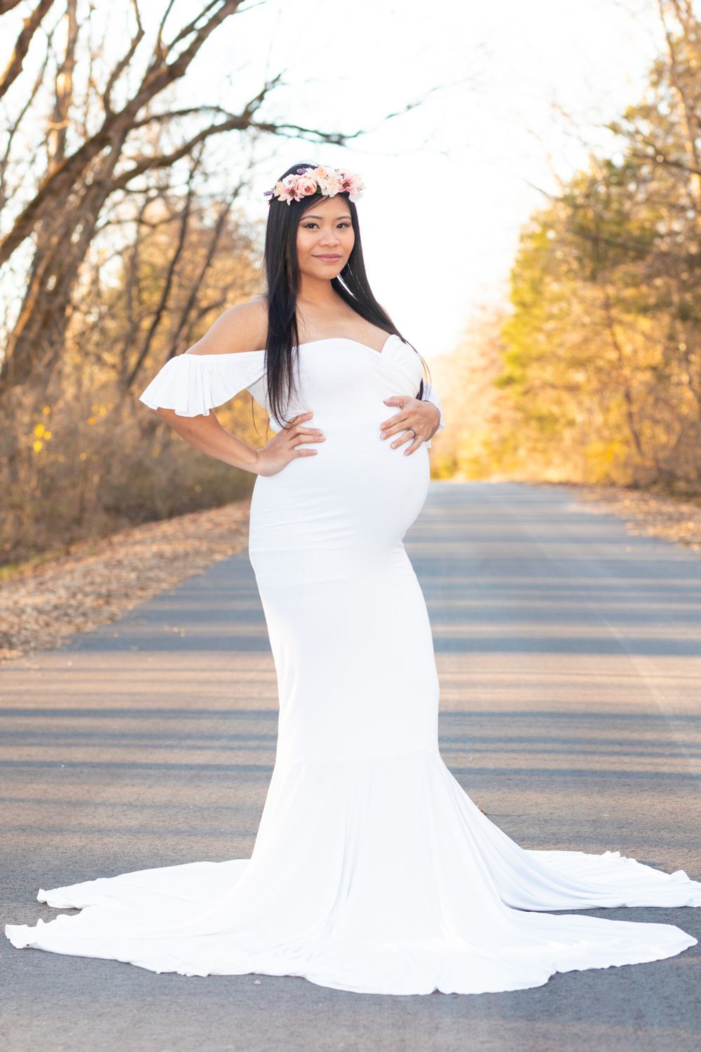 A soon-to-be mother wearing a white dress standing with her hands on her stomach looking at the camera.