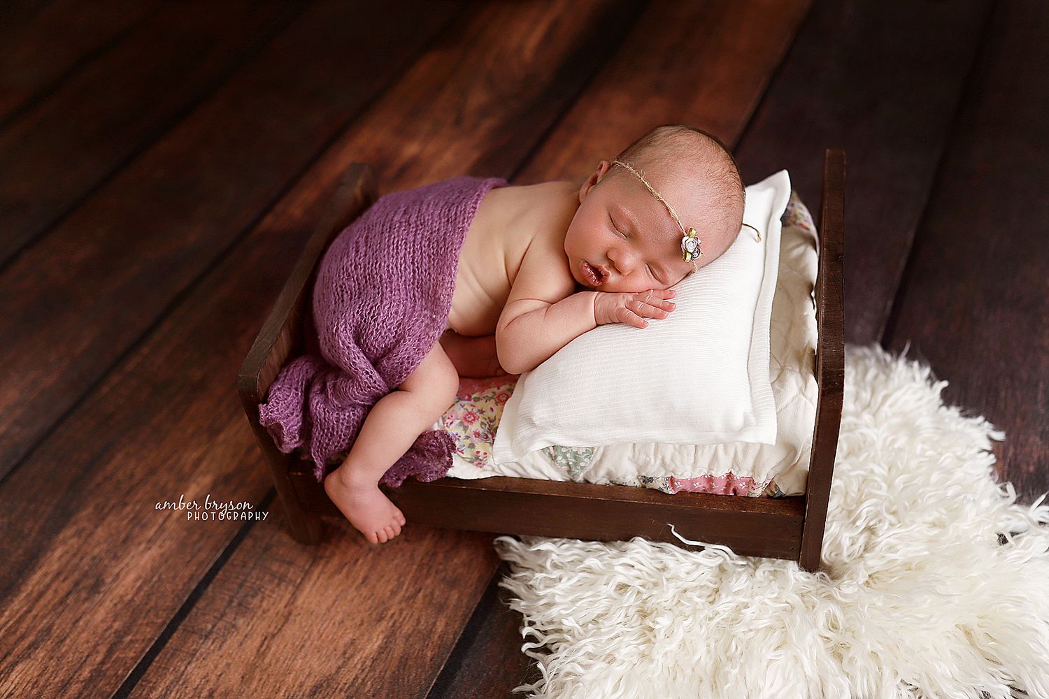 Sleeping newborn in purple wrap on white pillow against wooden backdrop.