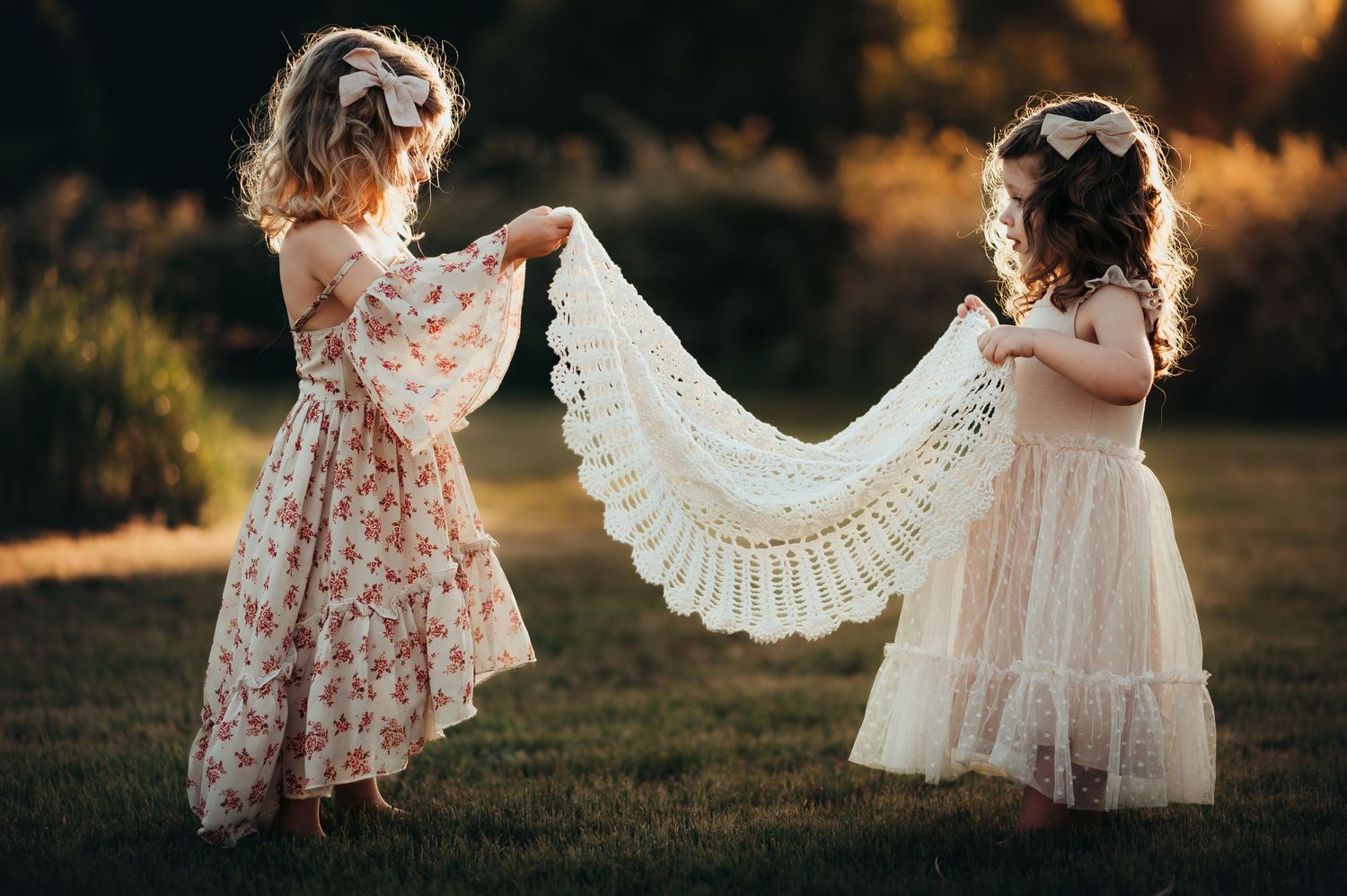 young girl cousins in pretty dresses and bows hold up a crochet blanket between them in a field at sunset.