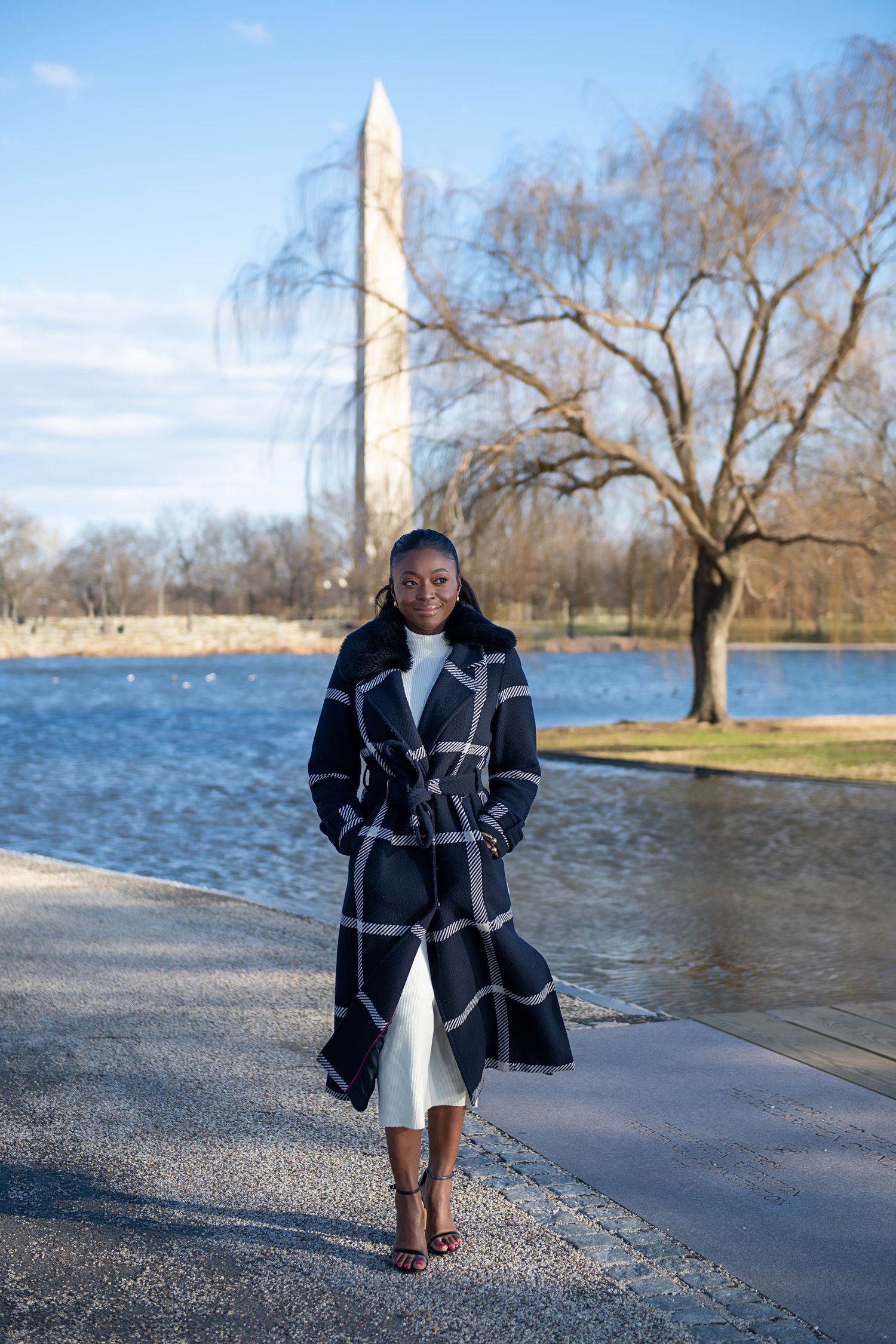 Person in plaid coat walking along reflecting pool with Washington Monument in background on sunny winter day.