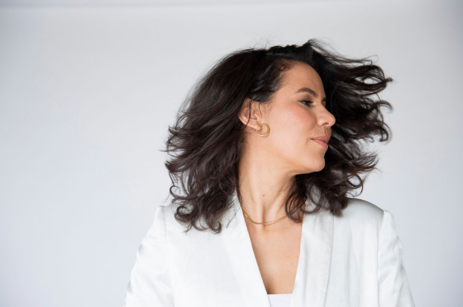 A person in a white blazer with wind-swept dark hair against a light background.