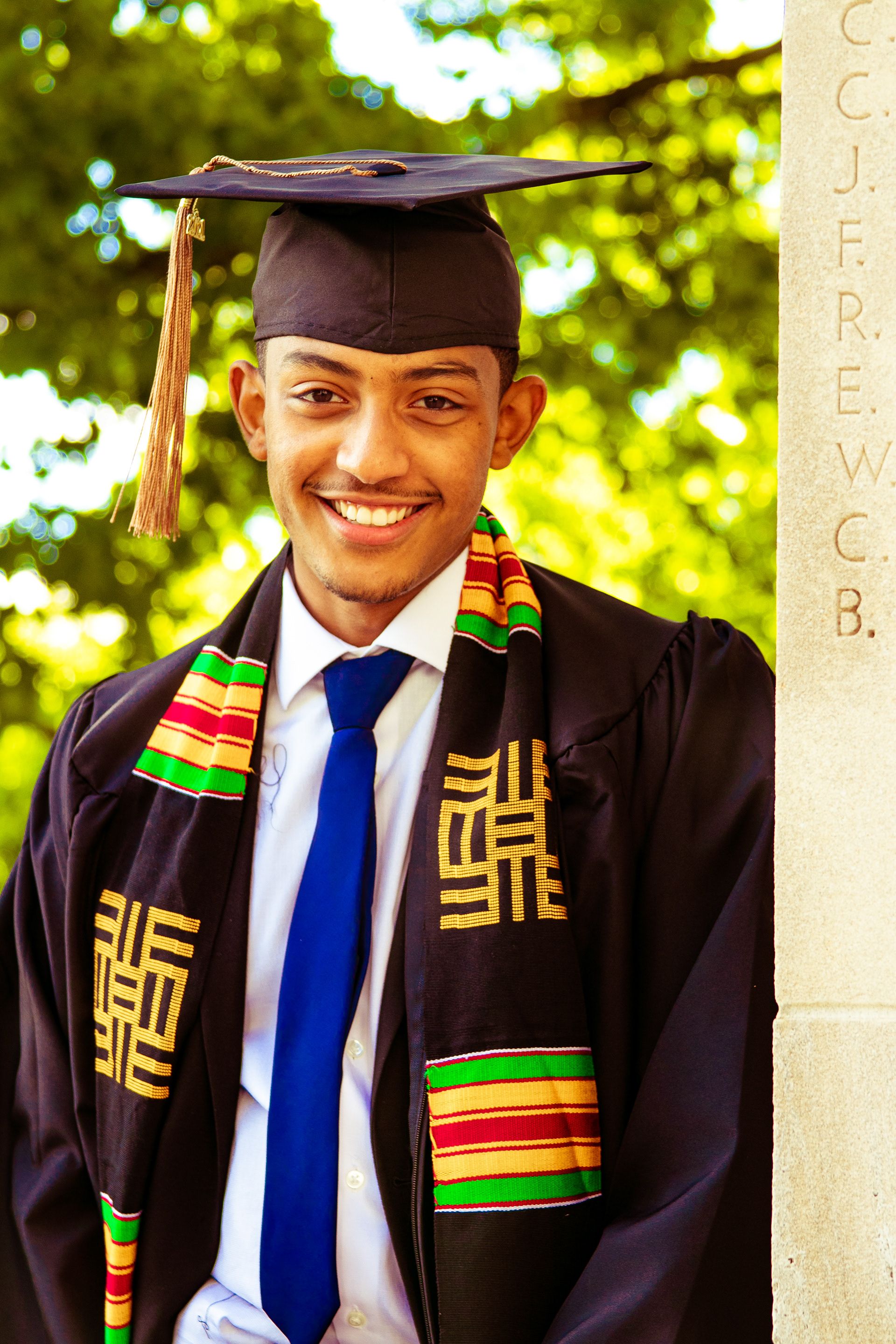Graduate in traditional black cap and gown with colorful kente stole smiling during graduation photo session.