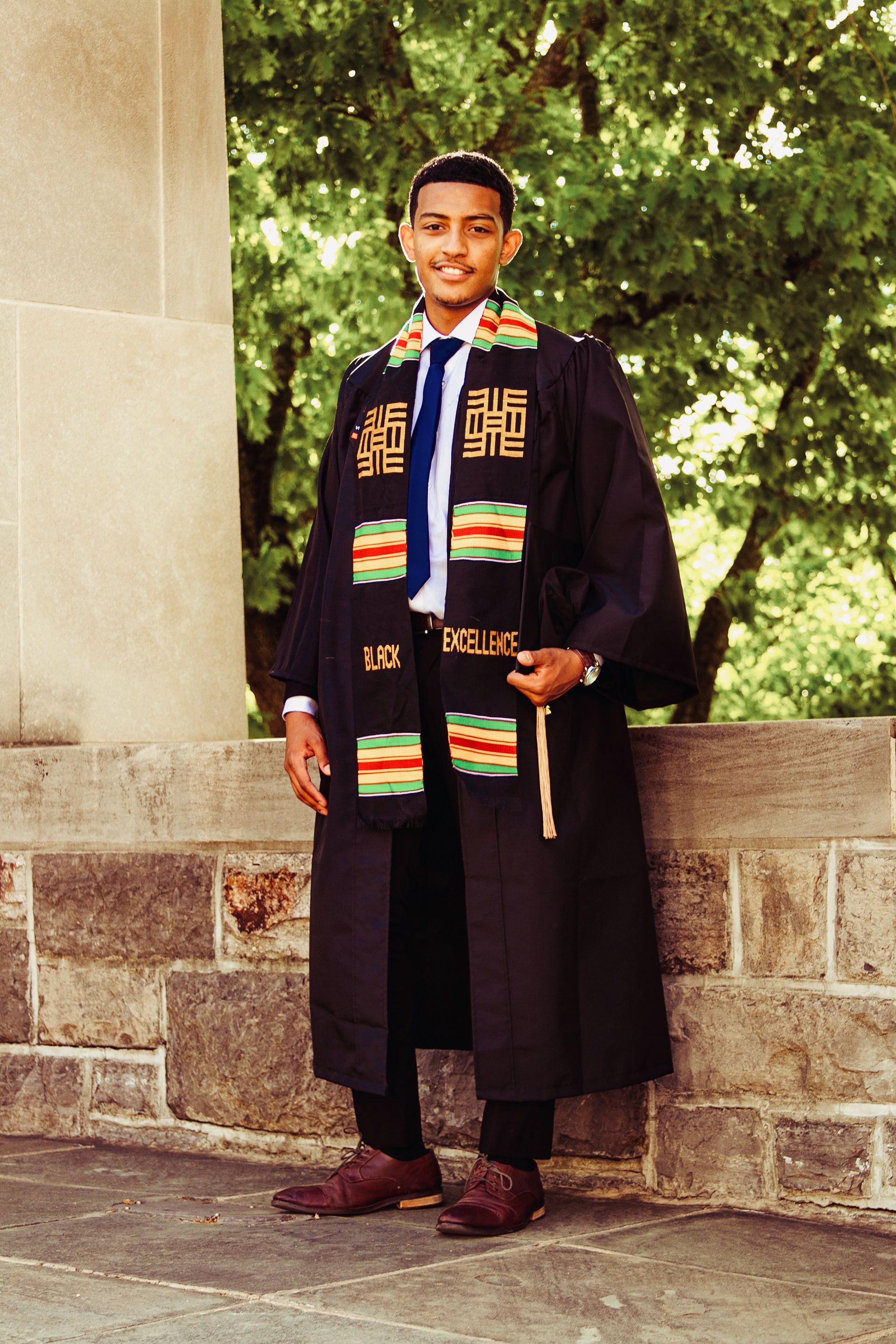 Graduates wearing traditional black robes with kente stoles sitting on stone steps outdoors.