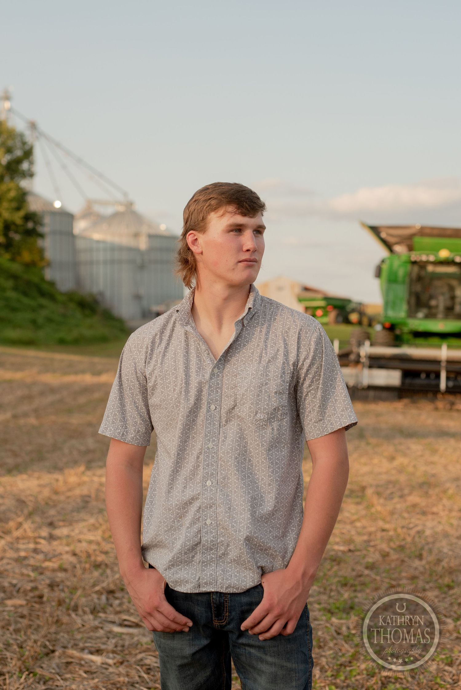 Portrait of a young person in casual attire standing in front of farm equipment and grain silos.