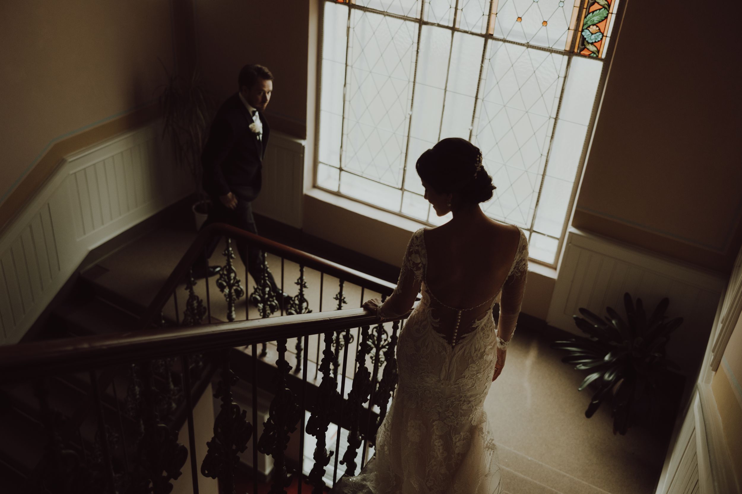 A dramatic wedding photo taken on a staircase with natural window light illuminating a bride and groom.