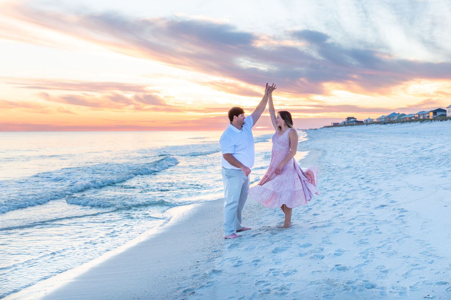Dancing couple on white sandy beach during vibrant sunset with coastal buildings in background.