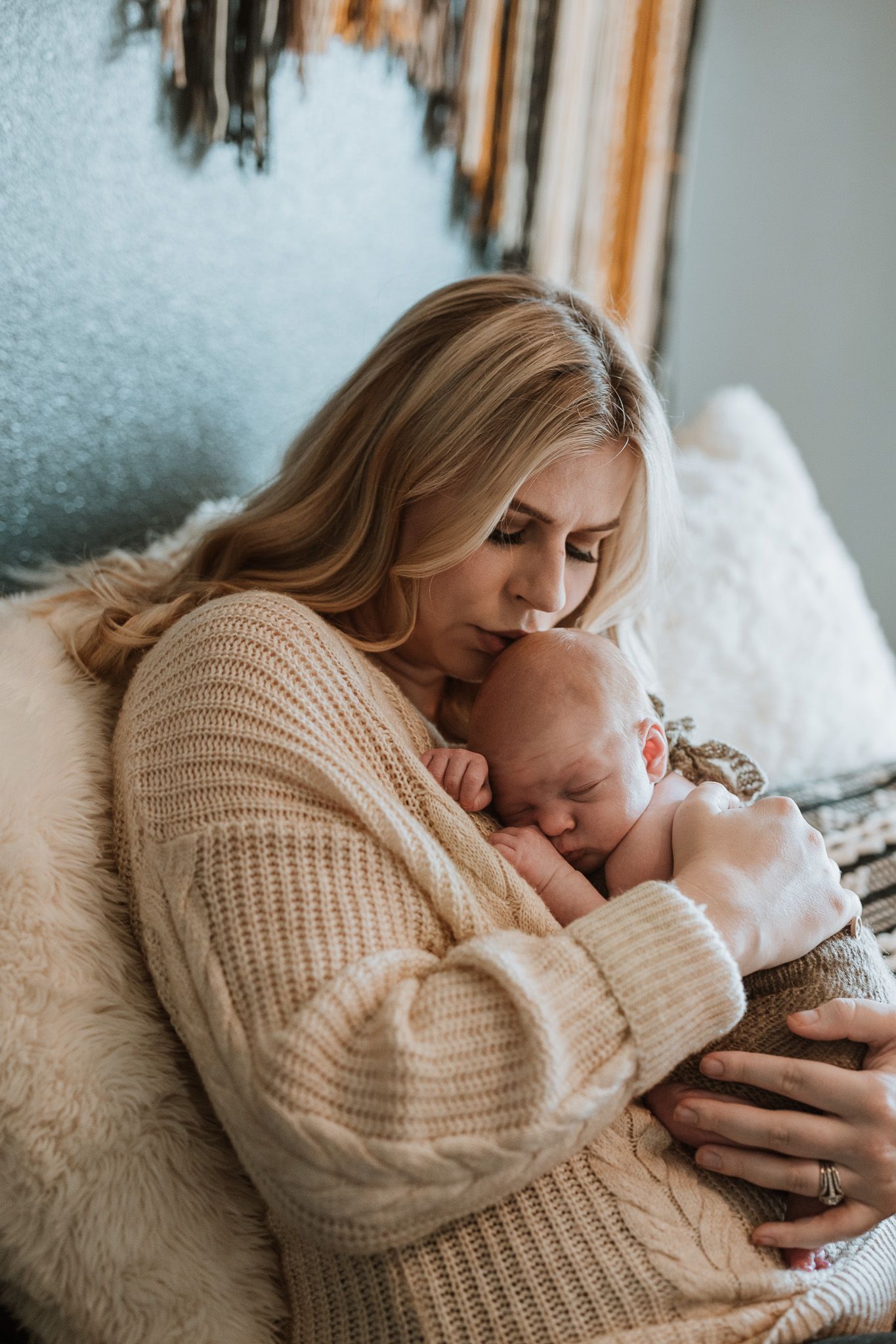 Tender moment as a parent in a cozy beige sweater cradles and kisses a sleeping newborn on soft white bedding.