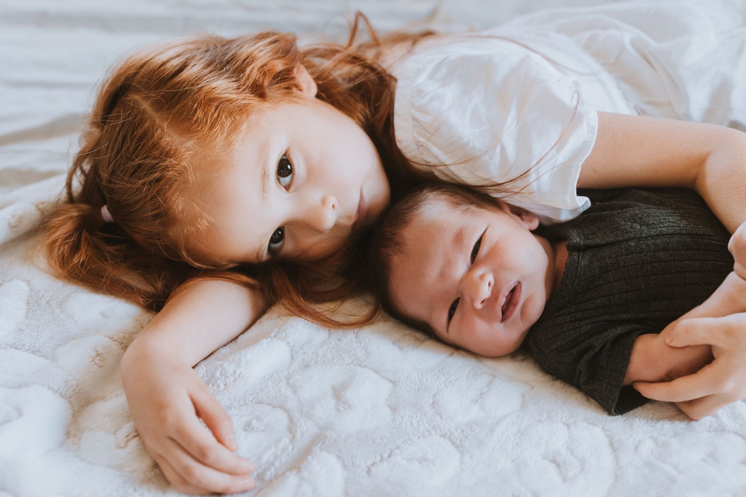 Two children lie together on white bedding during indoor lifestyle photoshoot.