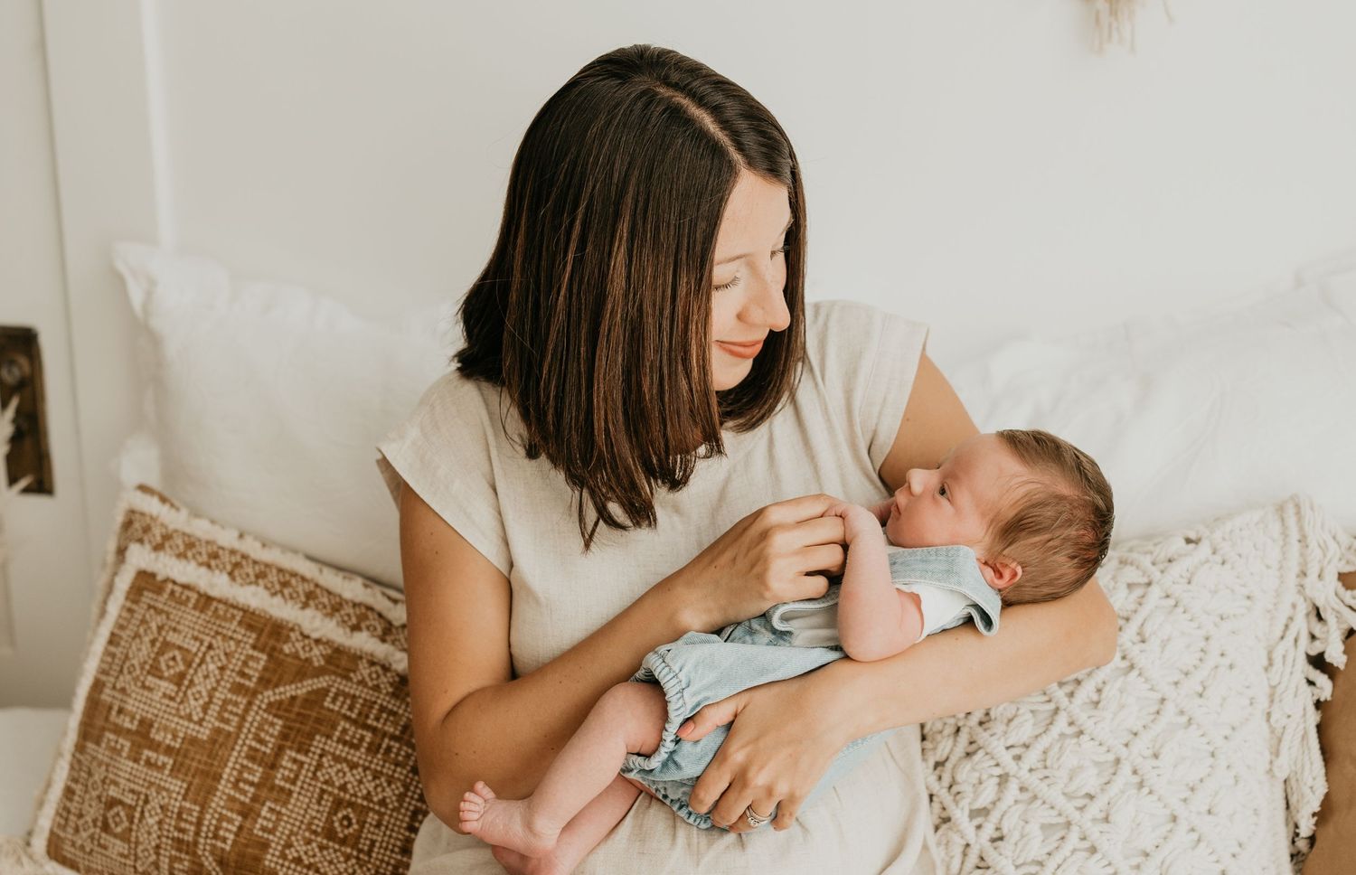Mother in white shirt bottle feeding baby on a cozy couch with macrame decor.