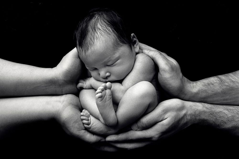 Dramatic black and white photo of newborn cradled in adult hands against dark background.