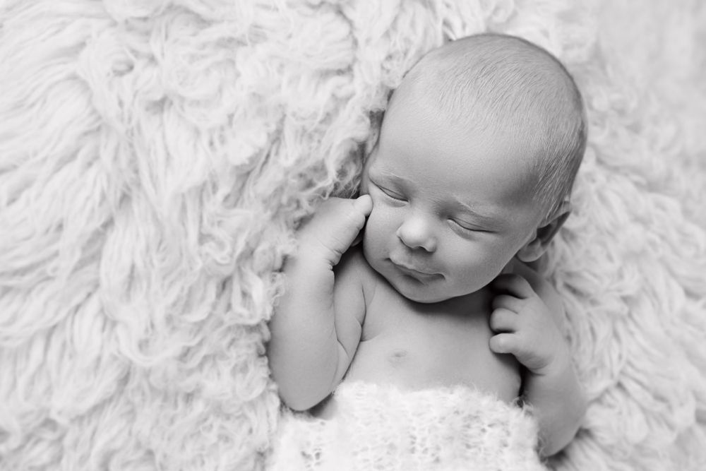 A sleeping newborn rests peacefully on plush white fur in monochrome.