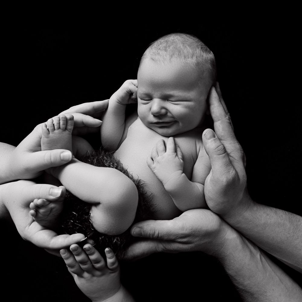 A laughing newborn baby supported by caring hands against black background.